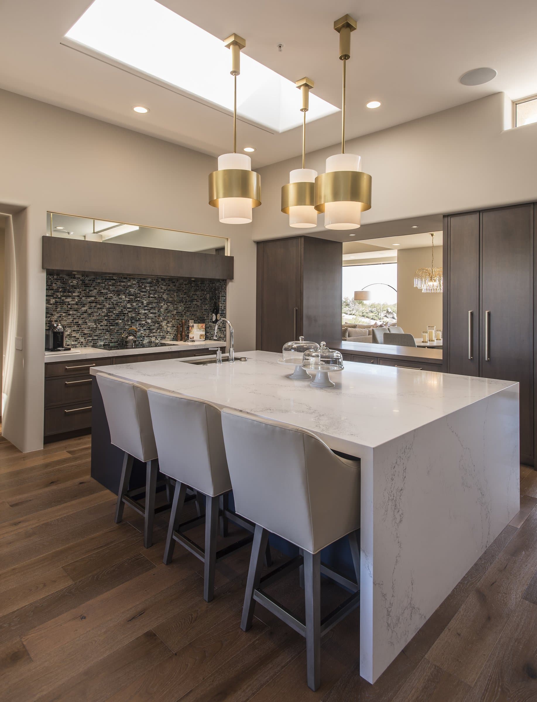 Luxury kitchen with a white marble waterfall island, grey bar stools, and gold-banded pendant lights under a skylight